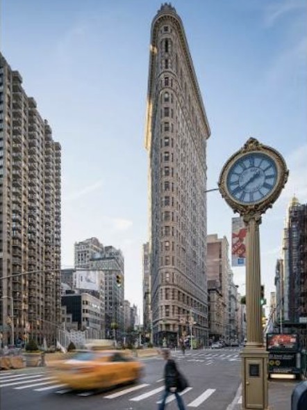 The Flatiron Building with the Fifth Avenue sidewalk clock in front and a taxi passing.
